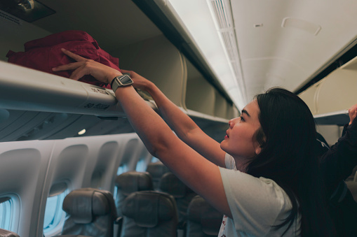 Female passenger storing handbag in overhead locker in airplane. Young woman in the cabin storing hand luggage in the overhead locke