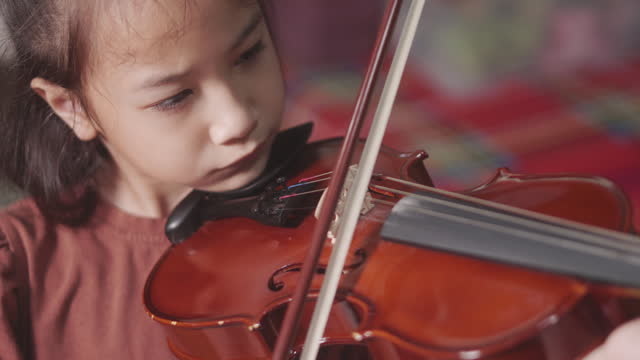 Close up 4K of cute Asian kid girl playing beautiful violin showing her face with determination for musical practice at home. Her small hands move bow across the wooden strings for beautiful song.