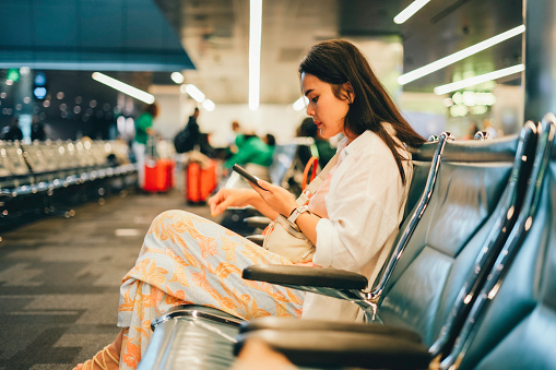 Young Asian woman using smartphone joyfully while waiting for night flight in airport terminal