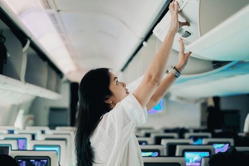 Female passenger storing handbag in overhead locker in airplane. Young woman in the cabin storing hand luggage in the overhead locke