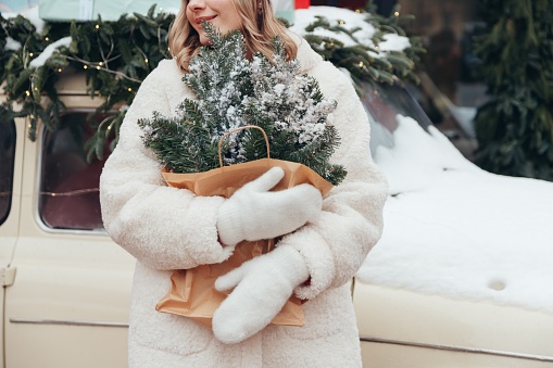 Woman with fir bouquet posing near beige retro car decorated with gift boxes for the New Year and Christmas.