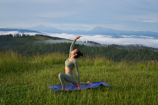 Relaxed woman in activewear practicing yoga on fresh air. Amazing view of summer mountains on background. Active lifestyles concept.