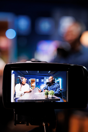 Selective focus on professional video camera screen recording bloggers streaming live internet radio using microphones from home studio. Black couple filming vlog for social media platform