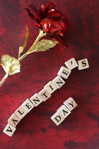 Stock photo showing elevated view of mottled red-black background with white letter dice spelling 'Valentine's Day' besides red and gold metallic, model rose, Valentine's Day greeting card sign.