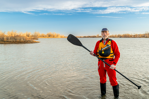 environmental portrait of a senior male paddler in a drysuit and life jacket holding stand up paddle on a shore of a lake