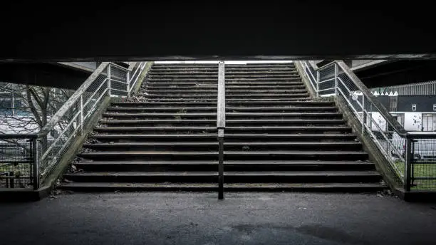 A set of darkly-lit stairs in a brutalist underpass at Crystal Palace National Sports stadium, strewn with litter and dead leaves
