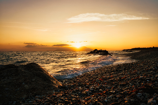 Beautiful sunrise in Greece. The sun rises behind the mountain and over the sea with waves. Waves roll over the pebble beach. The sun illuminates everything with warm shades of orange and yellow.