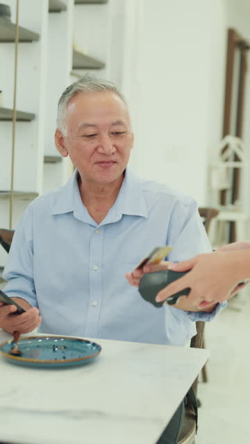 Elderly Asian male customer uses her smartphone to make a mobile payment at modern cafe, illustrating modern payment convenience. Small business owner. Vertical Screen.