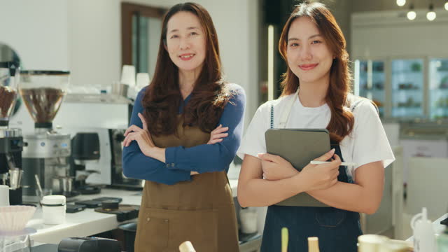 Two happy female entrepreneurs, a mentor and her trainee, pose with confidence at their successful and sustainable coffee shop. Small business owner.