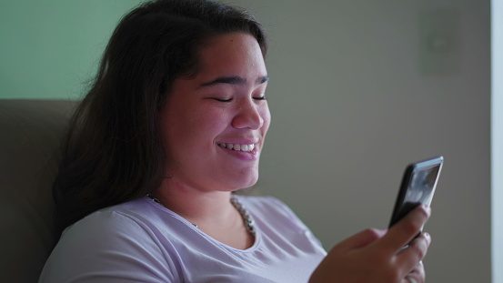 Happy Brazilian Teen Girl Reacts Positively to Online Content, Holding Phone. Candid scene of a Diverse Asian woman Smiling as She Reads Message