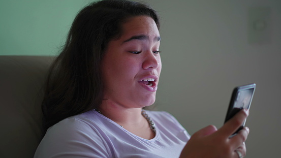 Happy Brazilian Teen Girl Reacts Positively to Online Content, Holding Phone. Candid scene of a Diverse Asian woman Smiling as She Reads Message