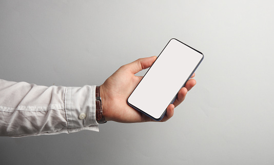 Man's hand in white shirt holds smartphone with white blank screen on gray background. Mockup, space for your information