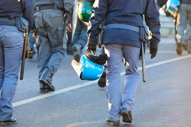 Group of heavily armed police officers or tactical operatives crouched in a defensive position amidst a chaotic environment. They are equipped with riot shields labeled \