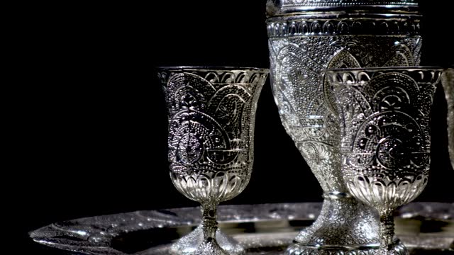 A side closeup of a silver ware set, with handmade cultural and traditional South Asian designs, rotating on an automatic display stand in black background and cinematic indoor light.