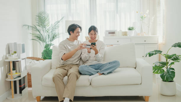 Young Asian couple having fun using smartphone devices shopping use credit card online payment together sit on sofa in living room at home. Lifestyle spend time in house.