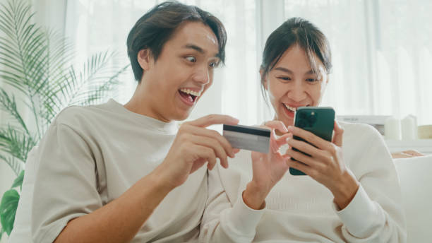 istock Young Asian couple having fun using smartphone devices shopping use credit card online payment together sit on sofa in living room at home. Lifestyle spend time in house. 1973069062