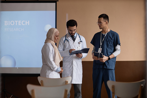 Medium full shot of Islamic men doctors looking through presentation notes in notebook of smiling female colleague wearing hijab while standing in conference room