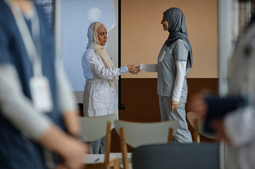 Medium full shot of two friendly Muslim women doctors wearing hijab handshaking in conference room