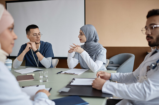 Islamic woman doctor wearing gray hijab speaking to male coworker sitting at table during conference