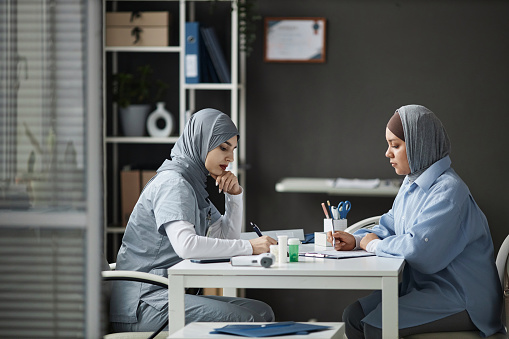 Side view of focused Muslim woman doctor and sick female patient wearing hijab filling in medical papers sitting at clinic desk