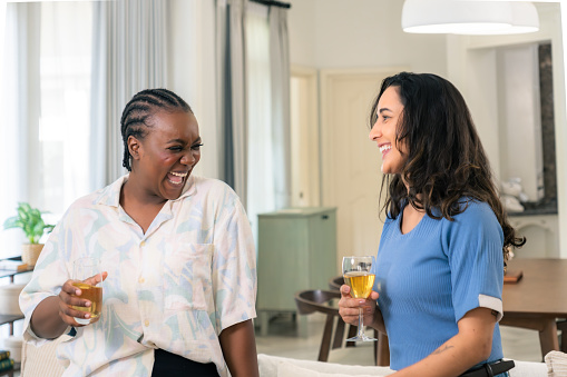 Cheerful African female laughing with Latin American woman while communicating together and drinking the celebratory toast at the living room on the occation of reunion for catching up each other with joy and happiness.