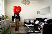 Girl hiding her face with a red heart-shaped balloon in the living room of the apartment