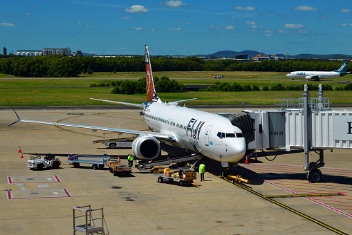Brisbane, Queensland, Australia: Fiji Airways Boeing 737 Max 8 - (registration DQ-FAB , MSN) 64307, ground services while parked at a passenger boarding bridge - in the background a Toll Aviation cargo Boeing 737-400 on a taxiway, Brisbane Airport (BNE).