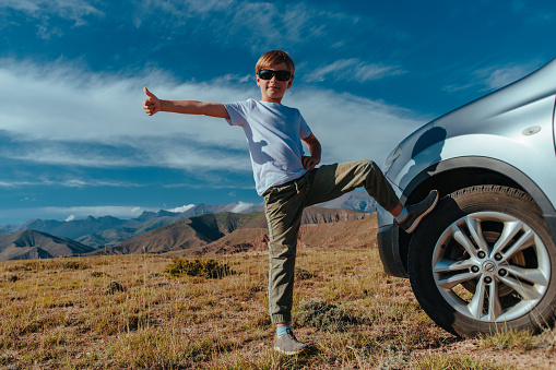 Happy boy in sunglasses stands by the car in the mountains and gives a thumbs up, travel concept