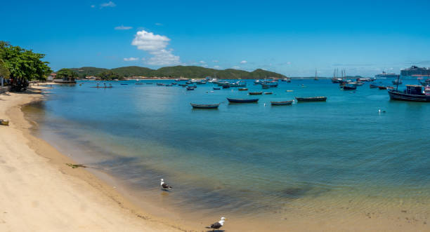 Drone photo of Armação dos Búzios, Brazil, showing the turquoise waters of the bay, small boats anchored offshore, a pier with a schooner, and a small island in the foreground. The image captures the coastal town and surrounding hills under a bright, clear sky.