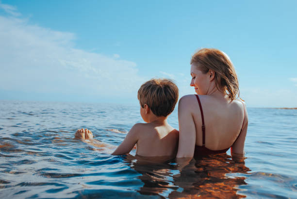 Mother and son sitting on seashore in water and looking away stock photo
