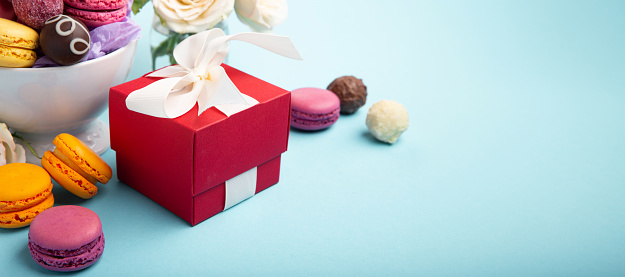 Sweet cookies macaroons, red gift box, rose flowers and chocolate candies on blue background. Presents concept for Mothers day, womans day, birthday, empty space for text message