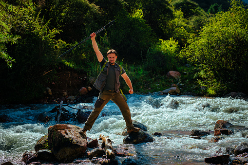 Happy hiker with trekking poles stands in the middle of a rushing mountain river