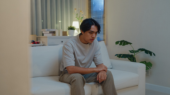 Sad depressed young Asian man sitting on sofa in living room at home night. Lifestyle spend time house concept.