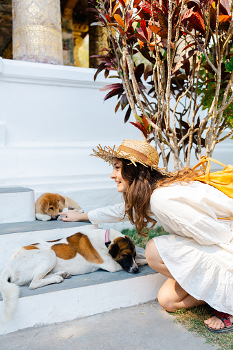 Cheerful woman in straw hat petting street dog laying on stairs during her vacation
