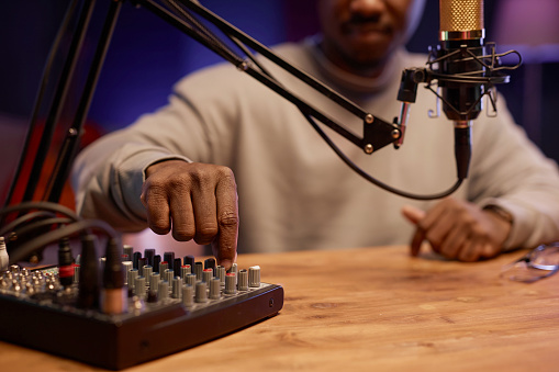 Cropped shot with focus on Black male hand switching controls on sound mixer board at desk with microphone in podcast studio