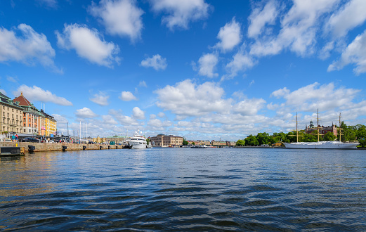 Stockholm, Sweden - July 15, 2023: Panoramic view on buildings and ships along embankment. Scandinavian architecture. Cityscape of Stockholm. Viewed from water in sunny summer day with a blue sky.
