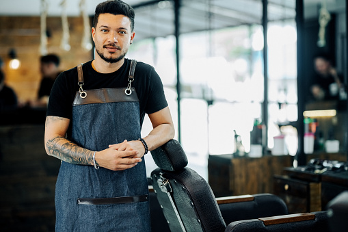 Portrait of a confident young male hairstylist in apron standing by chair in a salon