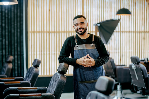 Portrait of a confident young male hairdressers with apron leaning to chair in a salon and looking at camera