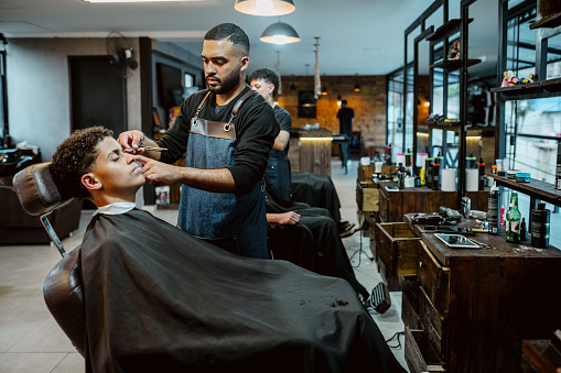 Hairdresser shaping eyebrows of man client using razor in barbershop