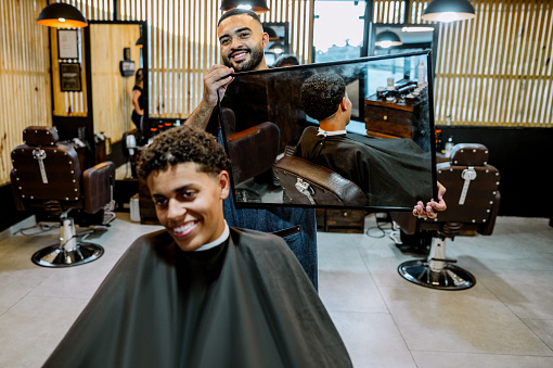 Barber showing the finished hairstyle with a mirror to the client sitting on the chair in barbershop