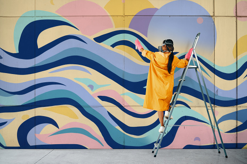 Female street artist painting colorful graffiti standing on a ladder. Modern art, urban concept.