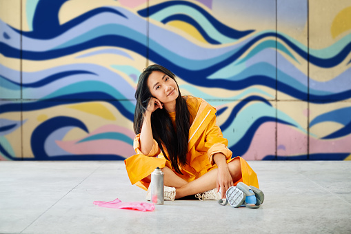 Female street artist rest sitting on the ground near the wall with her colorful paintings looking to camera. Street art concept