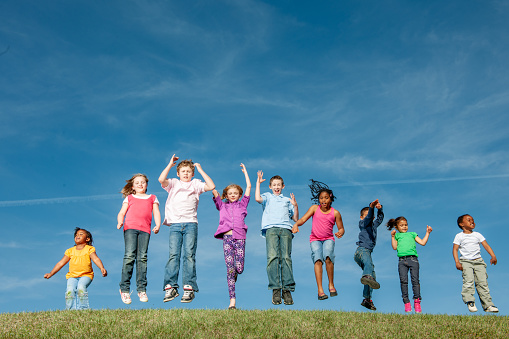 A large group of ethnically diverse school aged children jump side-by-side at the top of a grassy hill as they pose for a portrait.  They are each dressed casually and smiling as they are excited for summer.