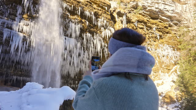 SLO MO Tilt Down Shot of Woman with Smartphone Photographing Peričnik Falls with Icicles Covered Cliff in Triglav National Park, Mojstrana