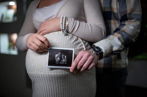 Close up of pregnant woman and her husband holding ultrasound image in front of her stomach - parenthood