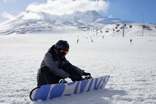 Girl or boy in ski helmet, sunscreen mask and balaclava sits on snow and fix the snowboard bindings against the backdrop of snow-covered mountain ski slope and a cloudy sky. Winter. Sport and travel content