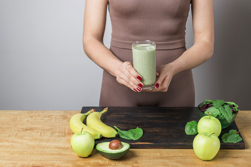 Unrecognizable athletic woman in sportswear holding glass of healthy green vegan smoothie. banana, spinach, apple and avocado on a table.