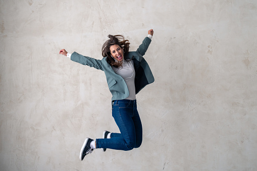 Full length of a jovial young woman in casual wear jumping in mid-air with arms up. Vertical shot. Isolated on white.