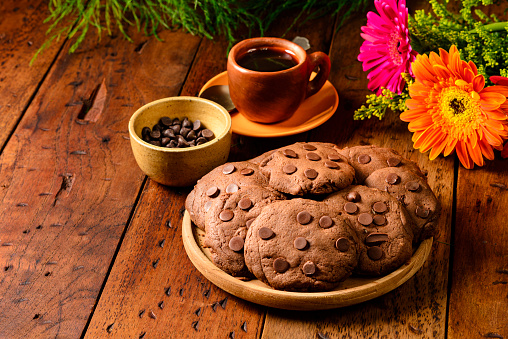 Front view of chocolate chip cookies made at home with gerbera flowers and cup of black coffee for a snack at home in the kitchen on a rustic wooden table