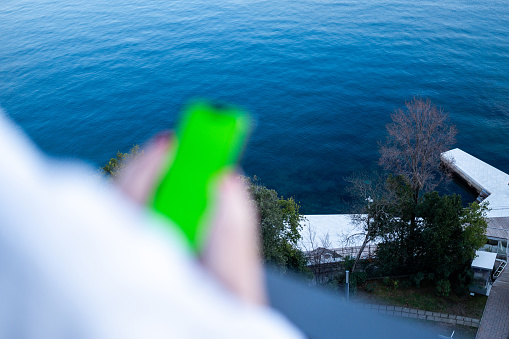 Young girl using mobile phone with greenscreen on balcony. Young girl using a phone at the seaside with a view using a phone with green screen - replacement screen.